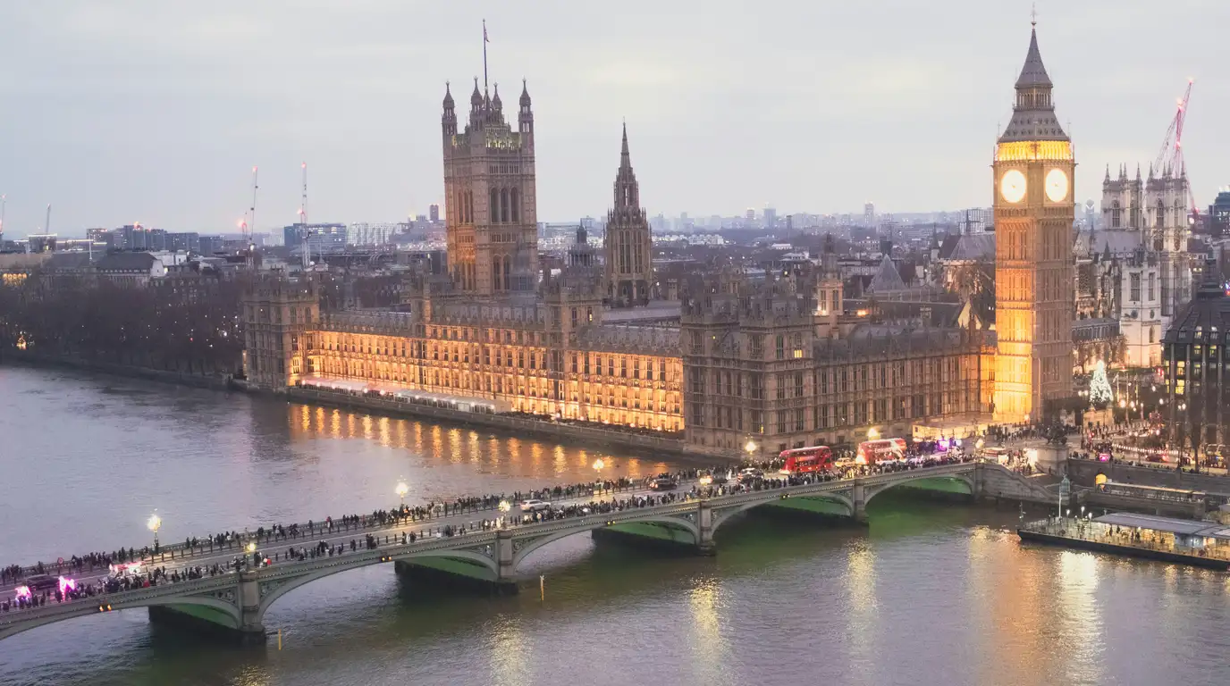 Riverside parliament buildings and a lit clock tower at dusk, with a crowded stone bridge and red buses crossing the water.