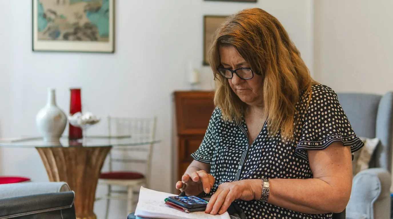 A woman in a living room working through a stack of papers, a pen in her hand and a phone resting on the pages.