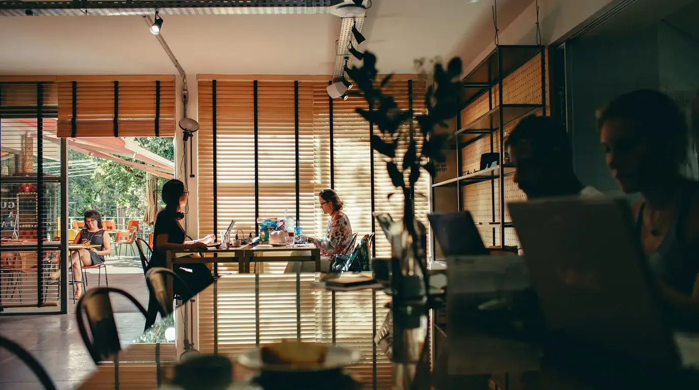A cafe work space with slatted blinds along the windows, several people at separate tables with laptops and notebooks.