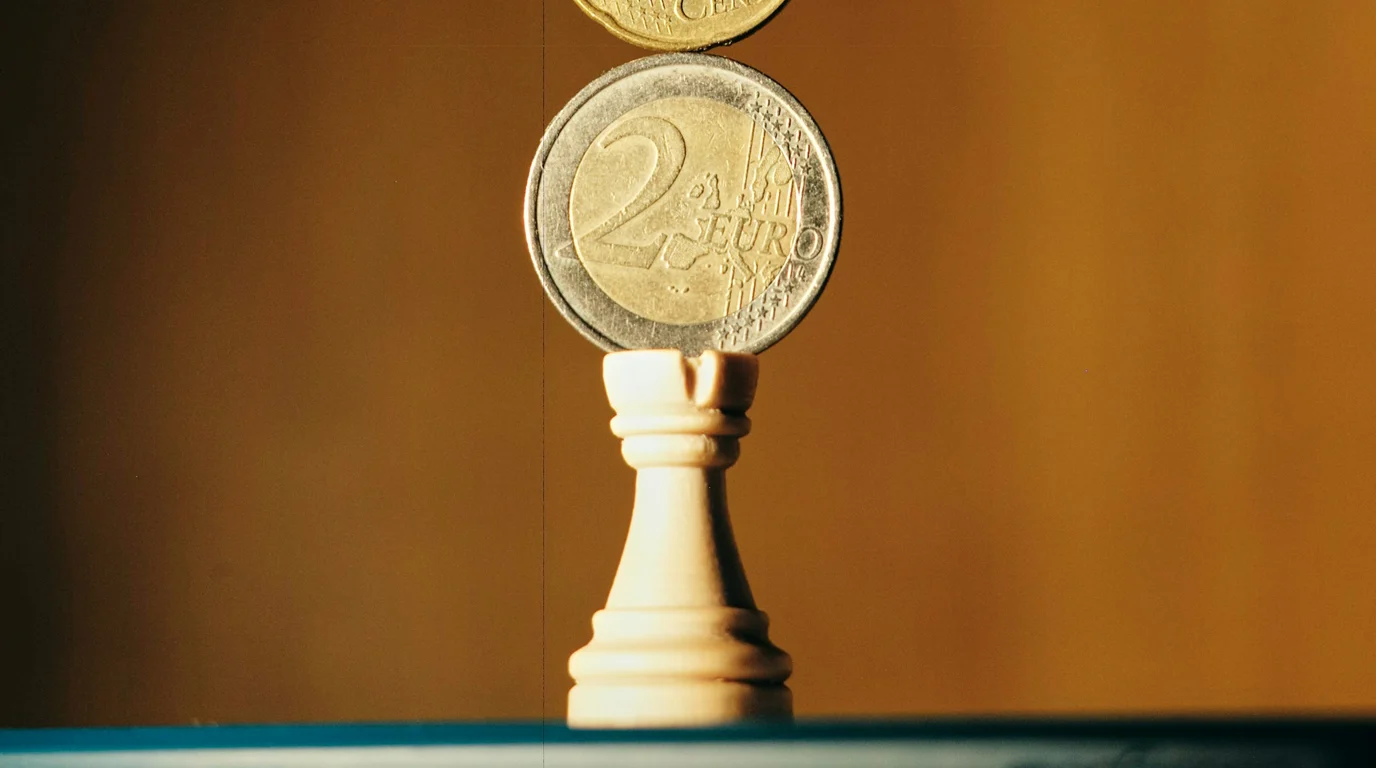 A two-euro coin balanced on the head of a white chess rook against a warm ochre backdrop, a second coin poised above it.