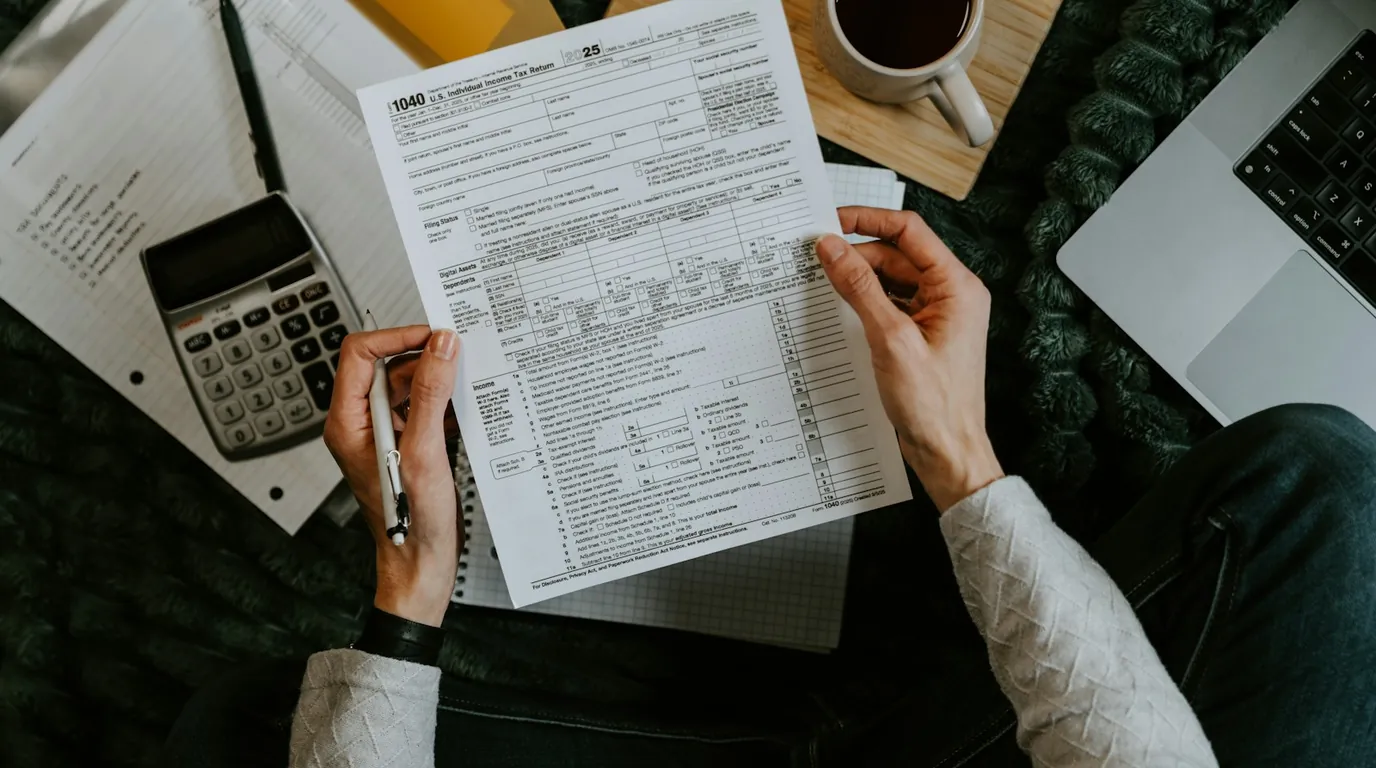 Hands holding a US tax form beside a calculator, a laptop and a cup of coffee.