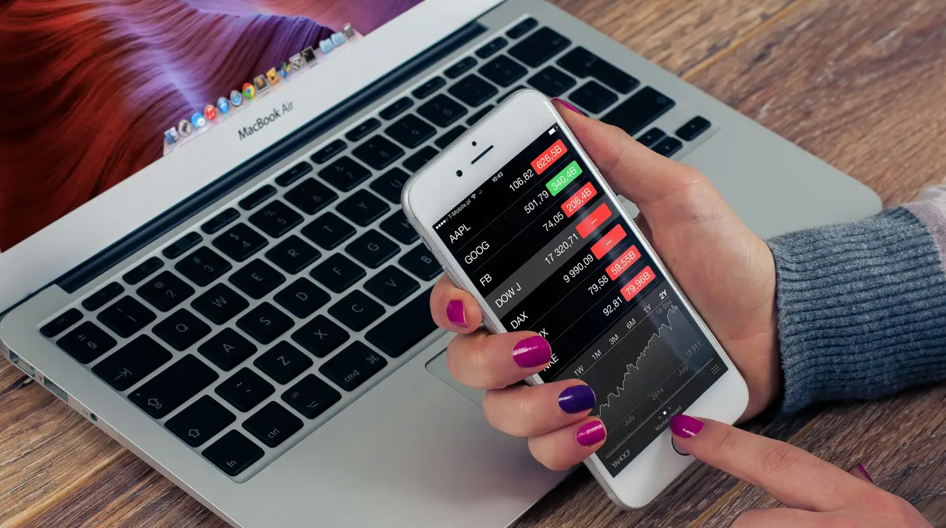 A manicured hand holds a smartphone showing a dark share-price list and line chart above an open laptop keyboard on a wooden desk.